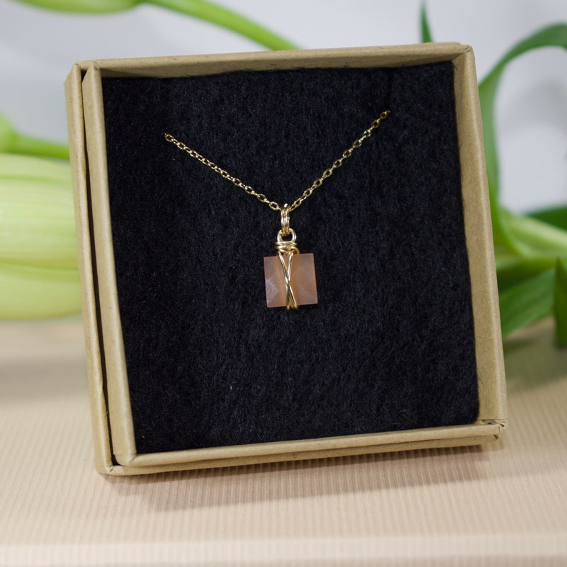 Pu&ograve; includere: Una collana color oro con un piccolo pendente quadrato color pesca. Il pendente &egrave; avvolto in filo d'oro e appeso a una delicata catena. La collana &egrave; presentata in una piccola scatola beige aperta con interno nero.