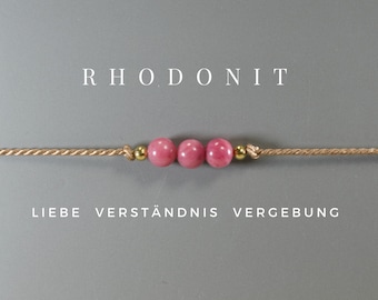 Può includere: Un semplice braccialetto con tre perline di rodonite rosa e dettagli dorati su un cordoncino beige. Il braccialetto ha le parole "Liebe, Verständnis, Vergebung" scritte sotto.
