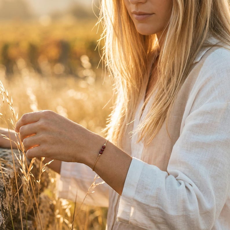 May include: A fine gold bracelet featuring three small, round, dark red gemstones. The bracelet is worn on a wrist, with a soft, blurred background of a field and a person wearing a white shirt.