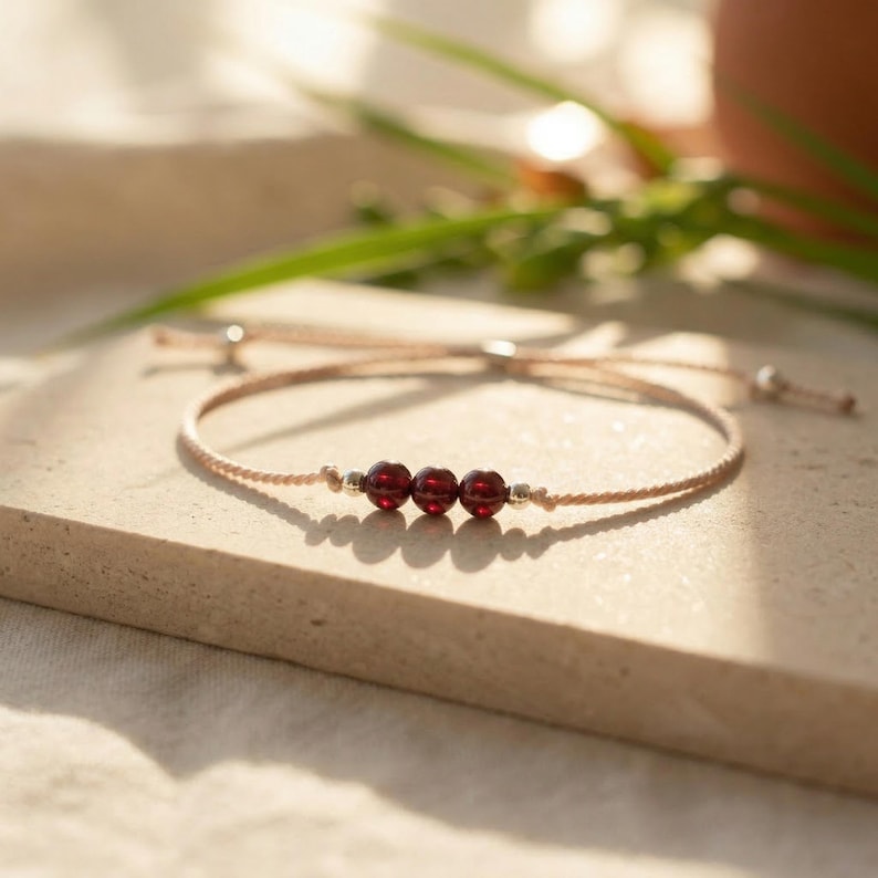 May include: A fine bracelet with three deep red gemstones. The stones are set between small silver-coloured beads on a thin, light brown cord. The bracelet is adjustable and looks handmade, suitable for everyday wear. The cord is approximately 15 cm in length.