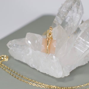 Pu&ograve; includere: Una collana color oro con un ciondolo quadrato color pesca. Il ciondolo &egrave; avvolto con filo metallico e poggia su una grande formazione di cristalli trasparenti. La catena della collana &egrave; visibile in primo piano.