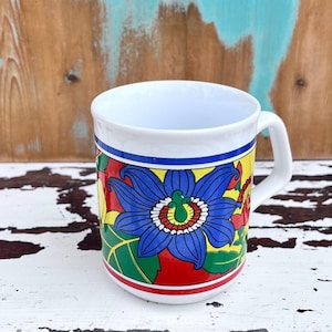 May include: White ceramic mug with a colorful floral design. The design features a large blue flower with red, yellow, and green accents.