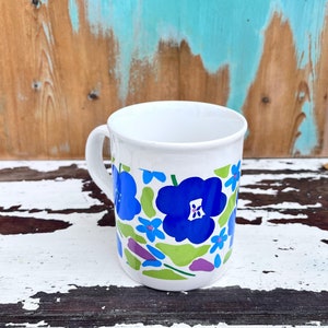 May include: White ceramic mug with a floral design featuring blue, green, and purple flowers. The mug has a handle and is sitting on a white and brown wooden surface.