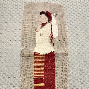 May include: A woven wall hanging featuring a folk art design of a person in traditional attire. The figure wears a white top, a red skirt, and a red head covering. The piece is made of natural-colored woven material.