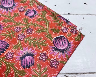 Op de afbeelding: Een levendige stof met een herhalend bloemenpatroon. Grote roze en paarse bloemen worden afgewisseld met kleinere roze bloemen en groene bladeren op een rode achtergrond. De stof is opgevouwen en onthult een wit oppervlak.