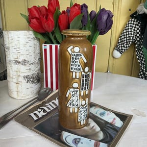 Może przedstawiać: Cylindryczny wazon ceramiczny z metalicznym brązowym szkliwem, ozdobiony czterema białymi wypukłymi figurami z czarnymi wzorami. Postacie są rozmieszczone pionowo na przedniej części wazonu, który posiada wąską szyjkę oraz płaską krawędź.
