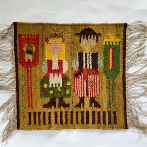 May include: Woven wall hanging featuring a folk art design. The image depicts two figures in traditional attire, flanked by decorative elements. The color palette includes gold, red, green, and brown, with fringed edges.