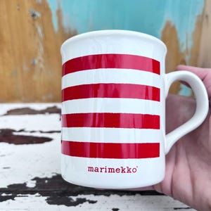 May include: White ceramic mug with bold red horizontal stripes. The mug features a curved handle and the word "marimekko" in red lettering near the base. The mug is a simple, yet stylish design.