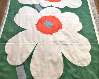 May include: Green fabric with a large floral pattern. The design features oversized white flowers with orange centers and blue accents. The stems are a light gray color. The fabric is likely used for home decor or textiles.
