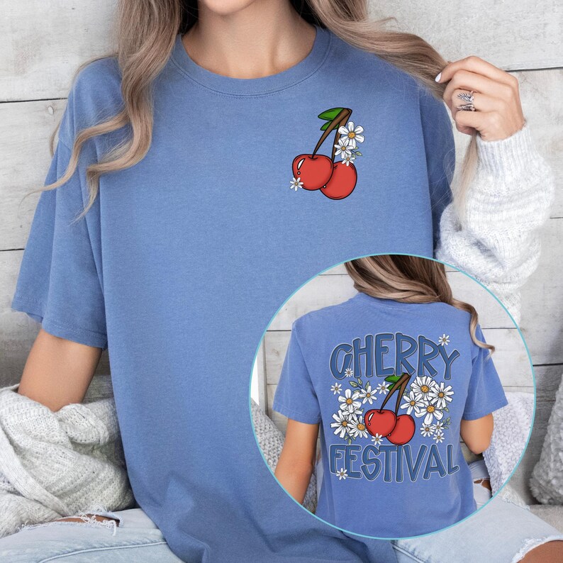 K&ouml;nnte beinhalten: Hellblaues T-Shirt mit einem Kirsch- und Blumendesign auf der Vorderseite. Auf der R&uuml;ckseite des Shirts steht "CHERRY FESTIVAL" mit Kirschen und G&auml;nsebl&uuml;mchen.