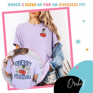 K&ouml;nnte beinhalten: Ein helllila T-Shirt mit einem Kirsch-Design auf der Vorder- und R&uuml;ckseite. Auf der R&uuml;ckseite des Shirts steht "Cherry Festival" mit einem Kirsch- und G&auml;nsebl&uuml;mchen-Design. Teil einer Bekleidungskollektion.