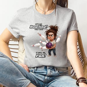 May include: A light gray t-shirt featuring a cartoon illustration of a person holding a purple bowling ball, surrounded by bowling pins. The text "Lets Go Bowling! They Said. It Will Be Fun!" is printed above and below the illustration.
