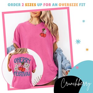 K&ouml;nnte beinhalten: Ein pinkfarbenes T-Shirt mit einem Kirsch- und G&auml;nsebl&uuml;mchen-Motiv. Auf der R&uuml;ckseite des Shirts steht "CHERRY FESTIVAL" in blauer Schrift. Das Shirt hat hochgekrempelte &Auml;rmel und eine lockere Passform.
