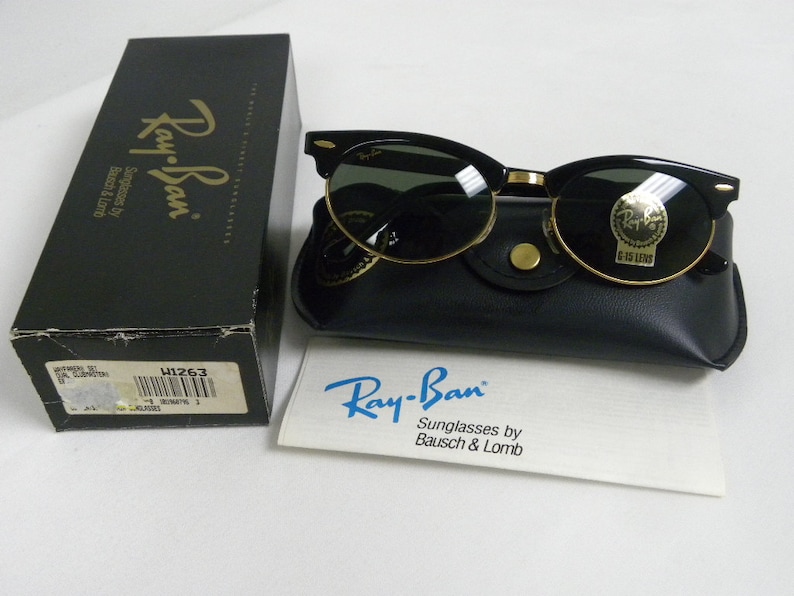 Puede incluir: Gafas de sol Ray-Ban negras con detalles dorados y lentes verdes. Las gafas de sol est&aacute;n en una funda de cuero negra con un bot&oacute;n dorado. La funda est&aacute; sobre una superficie blanca. Las gafas de sol est&aacute;n etiquetadas con "Ray-Ban Sunglasses by Bausch & Lomb".