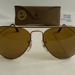 K&ouml;nnte beinhalten: Eine braune Aviator-Sonnenbrille mit goldenen Rahmen und braunen Gl&auml;sern. Die Sonnenbrille liegt auf einer wei&szlig;en Oberfl&auml;che.