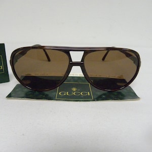 Puede incluir: Gafas de sol aviador de carey marrón con lentes marrones. Las gafas están sobre una tarjeta verde y blanca con la palabra "GUCCI" impresa en ella.