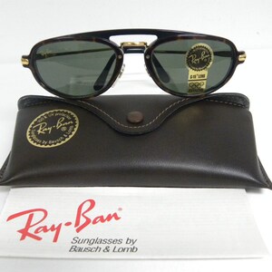 May include: Ray-Ban sunglasses with a tortoise shell frame and green lenses. The sunglasses rest on a brown leather case and a white paper with the Ray-Ban logo and text.