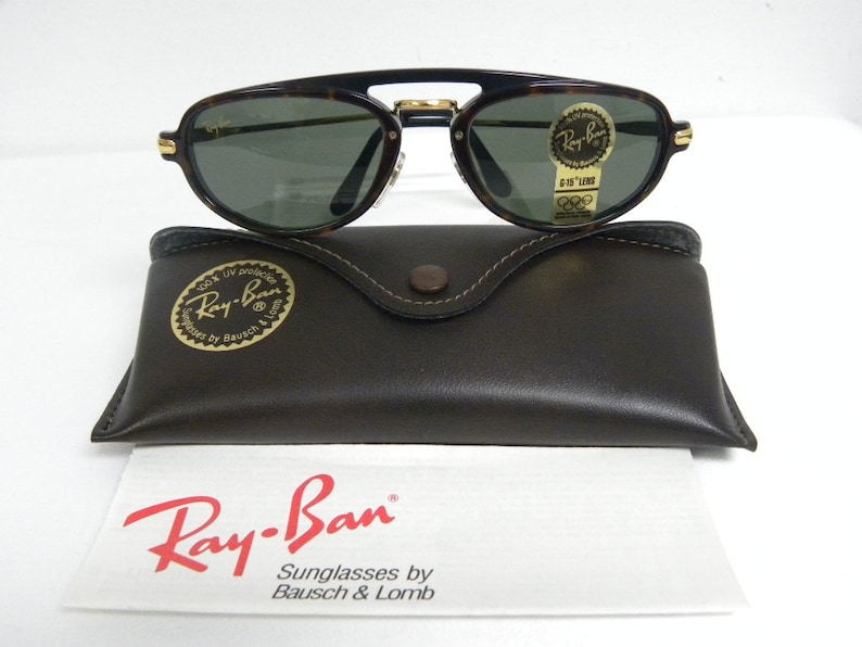 May include: Ray-Ban sunglasses with a tortoise shell frame and green lenses. The sunglasses rest on a brown leather case and a white paper with the Ray-Ban logo and text.