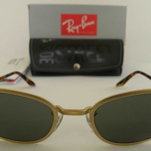 May include: Gold-tone Ray-Ban sunglasses with dark green lenses and a tortoise shell detail on the arms. The sunglasses are in focus and the background is blurred.