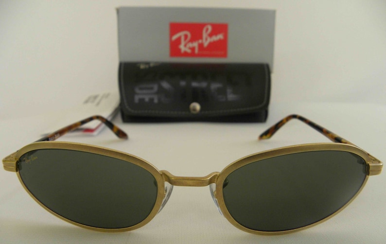May include: Gold-tone Ray-Ban sunglasses with dark green lenses and a tortoise shell detail on the arms. The sunglasses are in focus and the background is blurred.