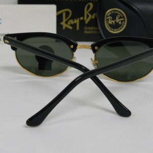 Puede incluir: Gafas de sol Ray-Ban negras con lentes verdes y detalles dorados. Las gafas est&aacute;n dobladas por la mitad y las patillas apuntan hacia la c&aacute;mara.