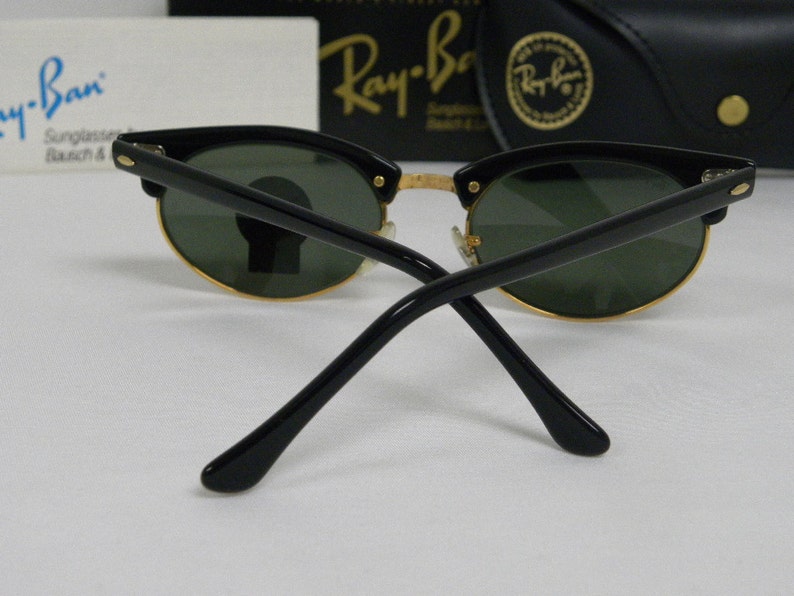 Puede incluir: Gafas de sol Ray-Ban negras con lentes verdes y detalles dorados. Las gafas est&aacute;n dobladas por la mitad y las patillas apuntan hacia la c&aacute;mara.