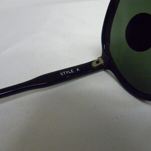 May include: Black sunglasses with green lenses and a silver hinge. The sunglasses have the text "STYLE A" printed on the arm.