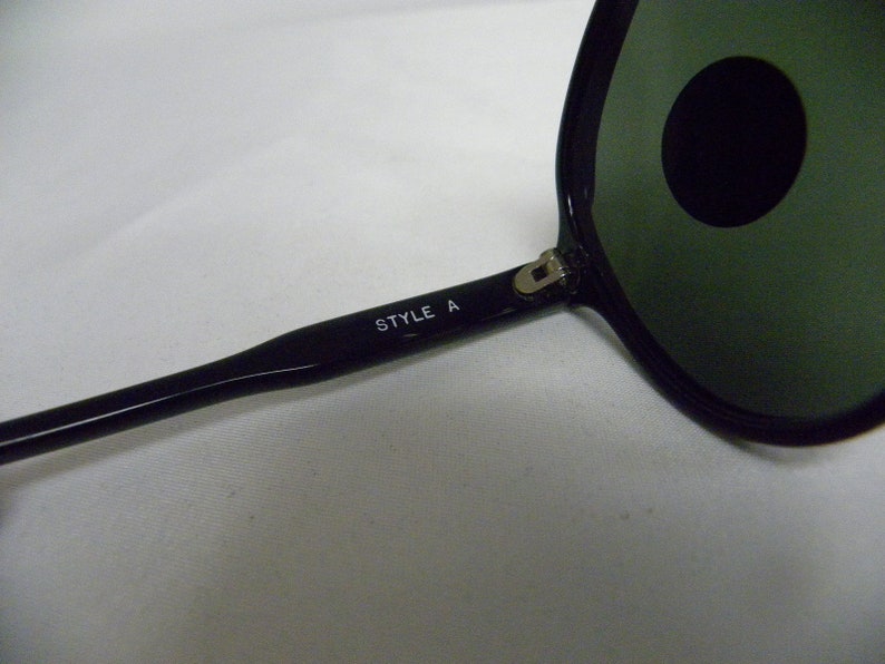 May include: Black sunglasses with green lenses and a silver hinge. The sunglasses have the text "STYLE A" printed on the arm.