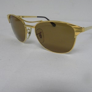 May include: Gold-tone aviator sunglasses with brown lenses. The sunglasses have a metal frame and a thin metal bar across the bridge of the nose.