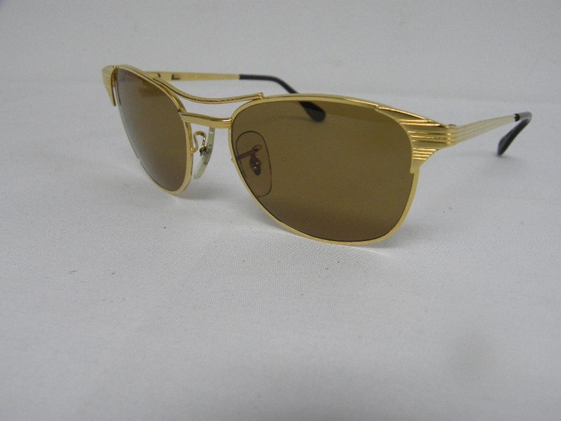 May include: Gold-tone aviator sunglasses with brown lenses. The sunglasses have a metal frame and a thin metal bar across the bridge of the nose.