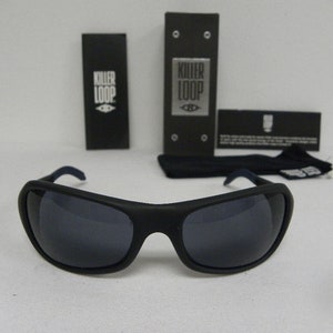 Op de afbeelding: Zwarte zonnebril met donkergrijze glazen. De zonnebril ligt op een wit oppervlak. De merknaam "Killer Loop" staat op de verpakking.
