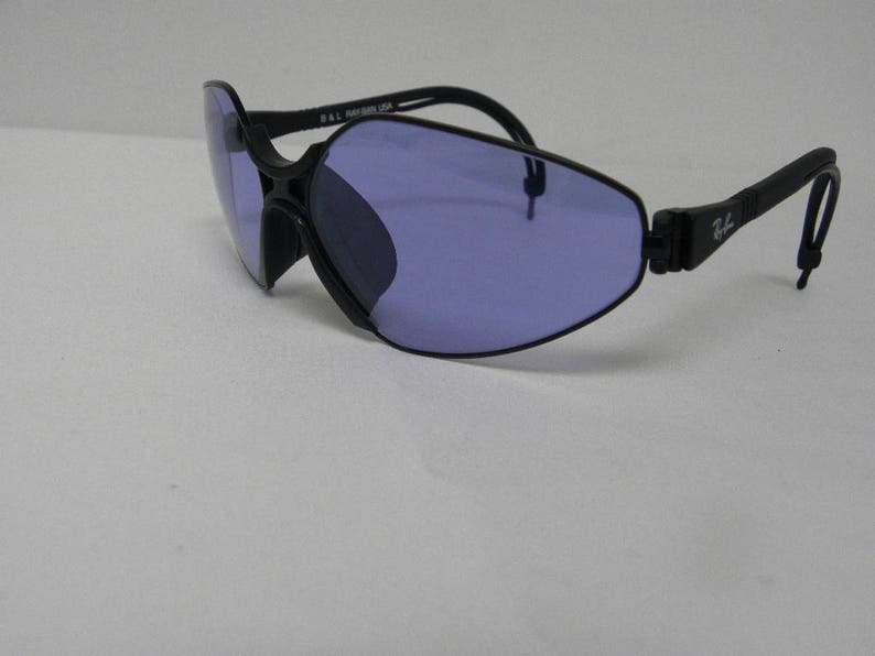 May include: Black sunglasses with a unique, angular frame and purple lenses. The sunglasses have a flexible, adjustable arm.