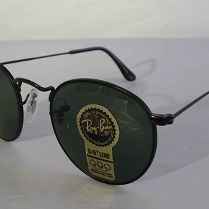K&ouml;nnte beinhalten: Schwarze Ray-Ban Sonnenbrille mit gr&uuml;nen Gl&auml;sern und einem goldenen Aufkleber mit der Aufschrift "100% UV protection Ray-Ban Sunglasses by Bausch & Lomb G-15 Lens" mit dem Olympischen Ringsymbol.