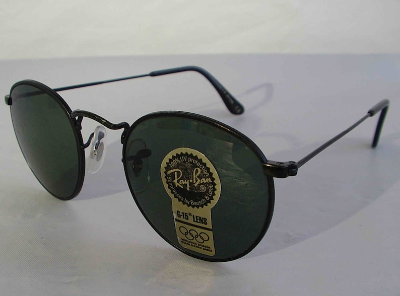 K&ouml;nnte beinhalten: Schwarze Ray-Ban Sonnenbrille mit gr&uuml;nen Gl&auml;sern und einem goldenen Aufkleber mit der Aufschrift "100% UV protection Ray-Ban Sunglasses by Bausch & Lomb G-15 Lens" mit dem Olympischen Ringsymbol.