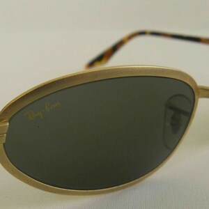 May include: Gold-tone oval-shaped sunglasses with dark green lenses and a tortoise shell temple. The sunglasses have the Ray-Ban logo on the left lens.