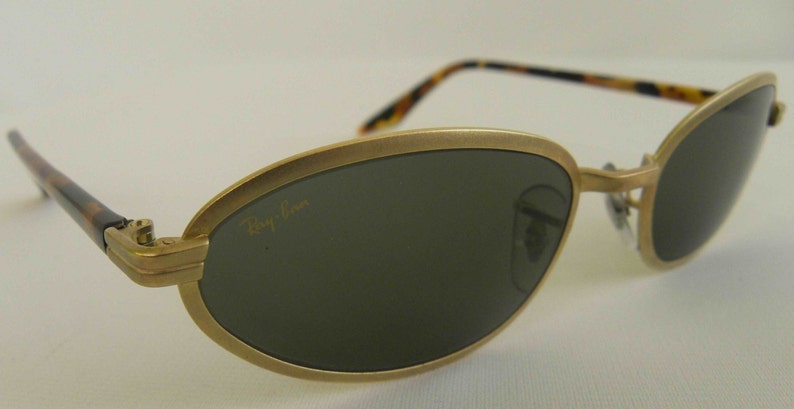 May include: Gold-tone oval-shaped sunglasses with dark green lenses and a tortoise shell temple. The sunglasses have the Ray-Ban logo on the left lens.