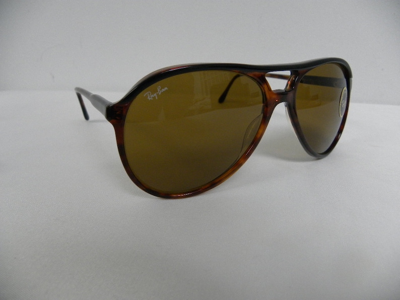 Puede incluir: Gafas de sol aviador de carey con lentes marrones. Las gafas tienen una barra de metal sobre el puente de la nariz y el logotipo de Ray-Ban es visible en la lente derecha.