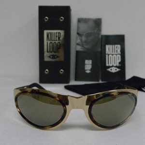 Op de afbeelding: Goudkleurige zonnebril met bruine glazen en een schildpadpatroon op de randen. De zonnebril wordt geleverd met een zwarte etui en twee zwarte kaarten, alle met de tekst "KILLER LOOP". Een zwart zakje is ook inbegrepen.