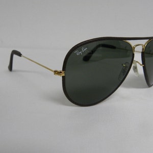 May include: Brown leather aviator sunglasses with gold frames and dark green lenses. The sunglasses have the Ray-Ban logo on the left lens.