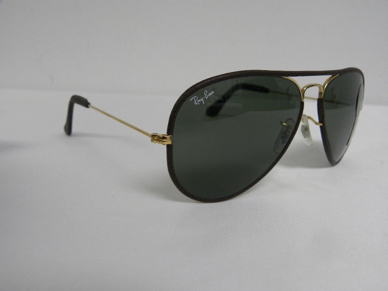 May include: Brown leather aviator sunglasses with gold frames and dark green lenses. The sunglasses have the Ray-Ban logo on the left lens.