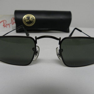 May include: Black Ray-Ban sunglasses with square lenses and a thin metal frame. The sunglasses are sitting on a white surface.