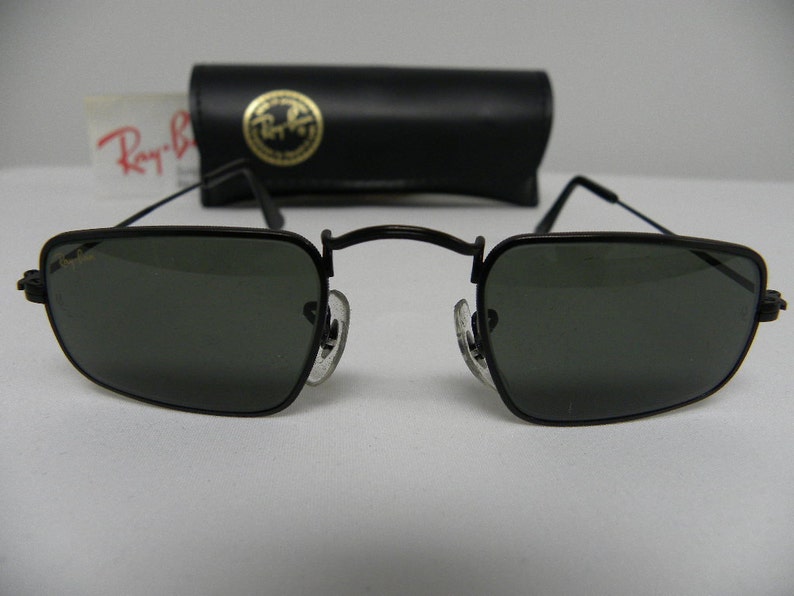 May include: Black Ray-Ban sunglasses with square lenses and a thin metal frame. The sunglasses are sitting on a white surface.