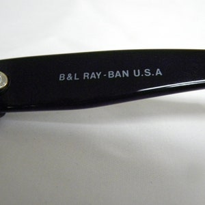 Puede incluir: Primer plano del brazo de unas gafas de sol negras con el texto "B&L RAY-BAN U.S.A." impreso en el brazo.