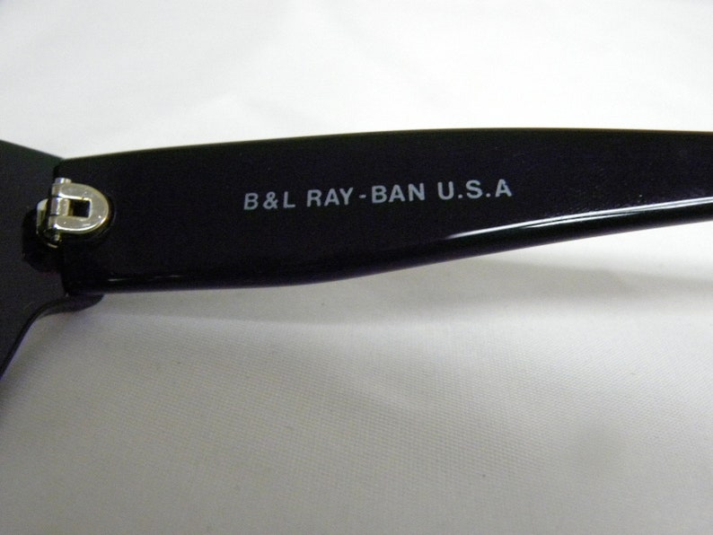 Puede incluir: Primer plano del brazo de unas gafas de sol negras con el texto "B&L RAY-BAN U.S.A." impreso en el brazo.