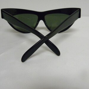 Puede incluir: Gafas de sol negras de ojo de gato con lentes verdes. Las gafas est&aacute;n dobladas por la mitad con las patillas cruzadas.