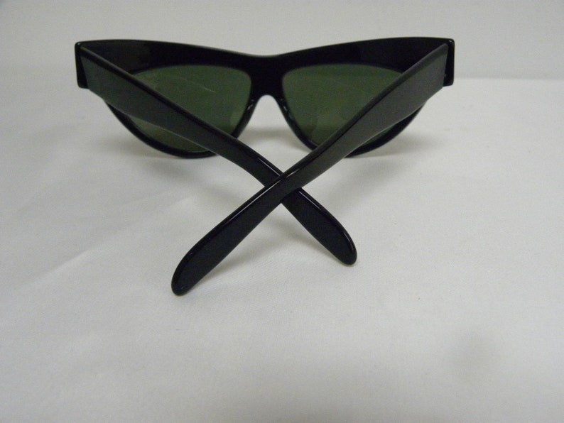 Puede incluir: Gafas de sol negras de ojo de gato con lentes verdes. Las gafas est&aacute;n dobladas por la mitad con las patillas cruzadas.