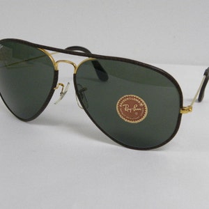 Może przedstawiać: Brązowe sk&oacute;rzane okulary przeciwsłoneczne typu aviator z pozłacanymi oprawkami i zielonymi szkłami. Okulary mają logo Ray-Ban na prawej soczewce.