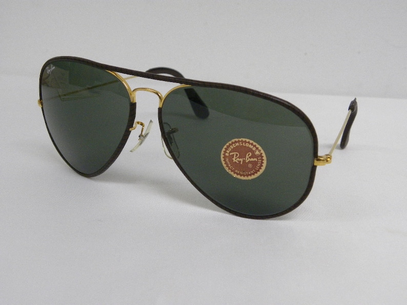 Może przedstawiać: Brązowe sk&oacute;rzane okulary przeciwsłoneczne typu aviator z pozłacanymi oprawkami i zielonymi szkłami. Okulary mają logo Ray-Ban na prawej soczewce.