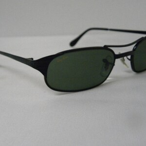 Puede incluir: Gafas de sol negras con montura de metal y lentes verdes. Las gafas tienen un dise&ntilde;o &uacute;nico con una barra de metal en la parte superior del marco.
