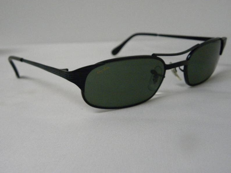 Puede incluir: Gafas de sol negras con montura de metal y lentes verdes. Las gafas tienen un dise&ntilde;o &uacute;nico con una barra de metal en la parte superior del marco.